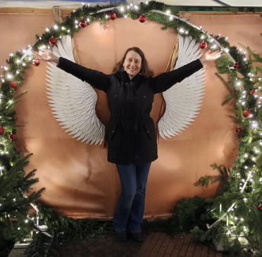 Elvira steht strahlend und mit ausgebreiteten Armen vor einer Wand mit Flügeln